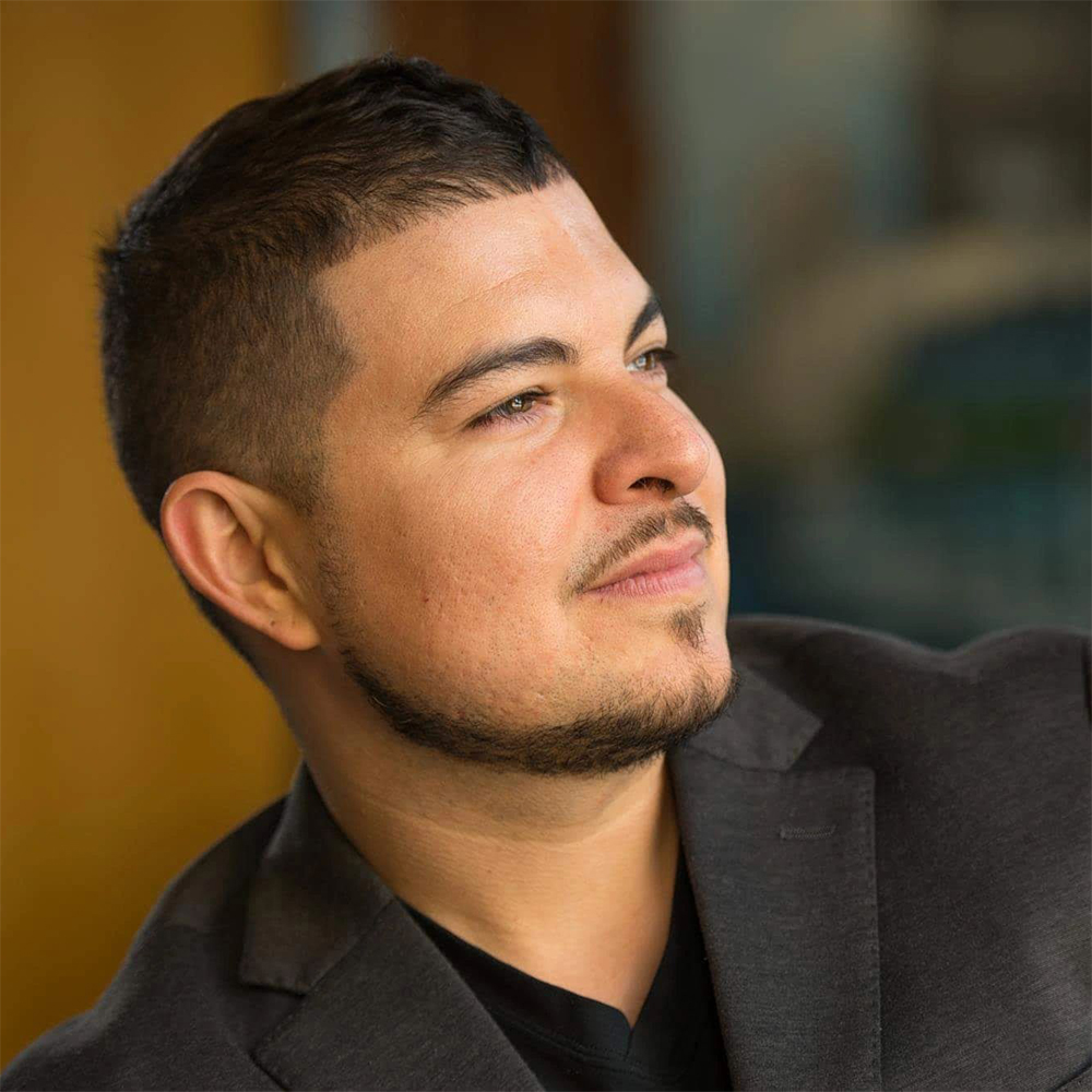 Portrait of Dr. Robert Rivera wearing a grey suit with a a black shirt looking off to the right